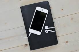 Celular com fones de ouvido sobre um caderno preto, em mesa de madeira