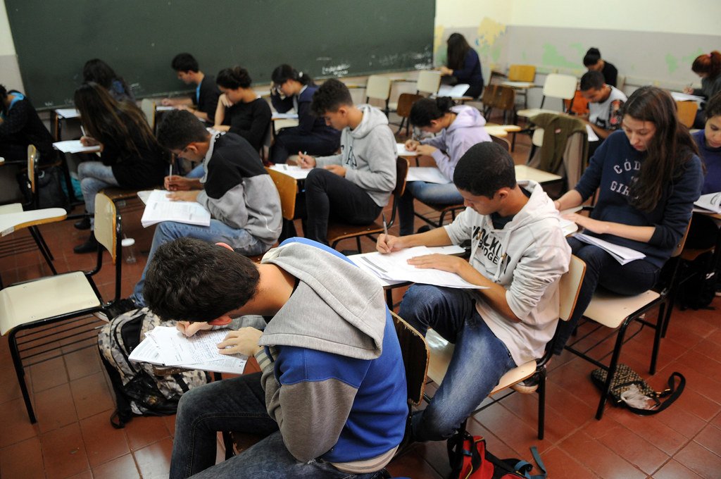 Candidatos sentados em carteiras escolares fazem uma prova escrita em sala de aula